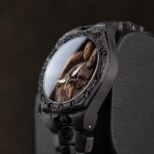 May include: A dark wooden wristwatch featuring a carved relief of two figures on the dial under a glass cover. The bezel has an intricate engraved pattern, and the watch case and band are constructed from dark, textured wood with visible grain.