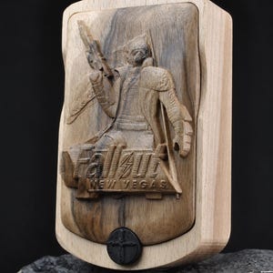 May include: A rectangular wooden box featuring a relief carving of a character holding a firearm, with the text Fallout New Vegas engraved below. The box has a light-coloured frame and a darker centre panel, with a small black circular emblem at the base.