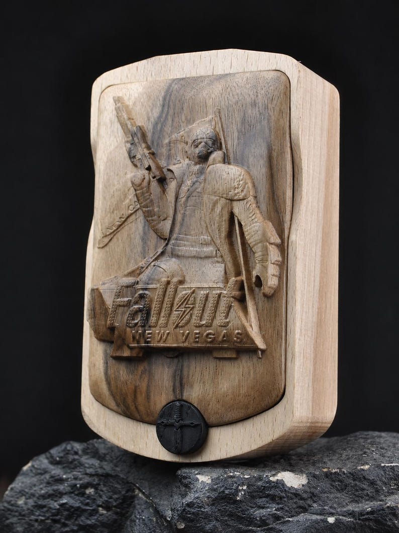 May include: A rectangular wooden box featuring a relief carving of a character holding a firearm, with the text Fallout New Vegas engraved below. The box has a light-coloured frame and a darker centre panel, with a small black circular emblem at the base.