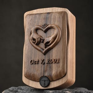 May include: A rectangular wooden box featuring a dark walnut center panel with a carved heart shape formed by two hands touching. Below the carving, the text Oct 7, 2001 is engraved. A small black circular button is positioned at the bottom center.