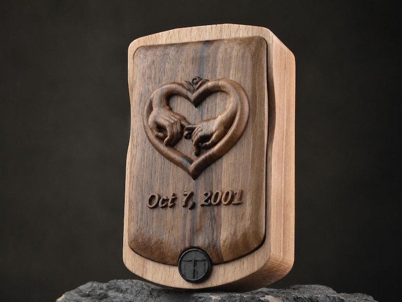 May include: A rectangular wooden box featuring a dark walnut center panel with a carved heart shape formed by two hands touching. Below the carving, the text Oct 7, 2001 is engraved. A small black circular button is positioned at the bottom center.