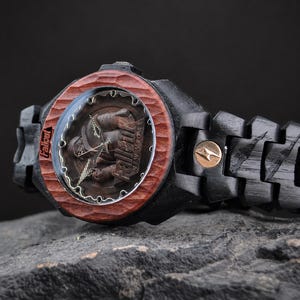 May include: A wristwatch with a black wooden link band and a carved reddish-brown wooden bezel. The watch face features a Fallout New Vegas graphic with the text Fallout and New Vegas visible. A small circular gold-toned emblem is on the band.