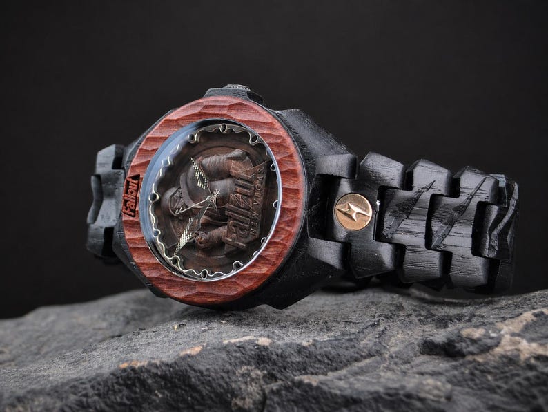May include: A wristwatch with a black wooden link band and a carved reddish-brown wooden bezel. The watch face features a Fallout New Vegas graphic with the text Fallout and New Vegas visible. A small circular gold-toned emblem is on the band.