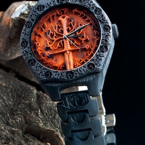 Puede incluir: Un reloj de pulsera de madera negra con una banda negra. La esfera del reloj presenta un árbol tallado con una espada y la inscripción "Un anillo para gobernarlos a todos, Un anillo para encontrarlos, Un anillo para atraerlos a todos y en las tinieblas atarlos."