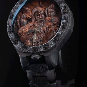 May include: Black wristwatch with a detailed, carved wooden face featuring a bearded figure with birds. The watch has a black band and casing with intricate patterns around the face. The watch face is approximately 1.75 inches in diameter.