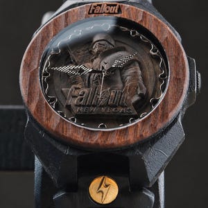 May include: A black wooden wristwatch featuring a dark wood bezel and a dial with a carved Fallout New Vegas character relief. The watch face displays the Fallout logo and text, with metallic hands and a small gold circular emblem on the strap.