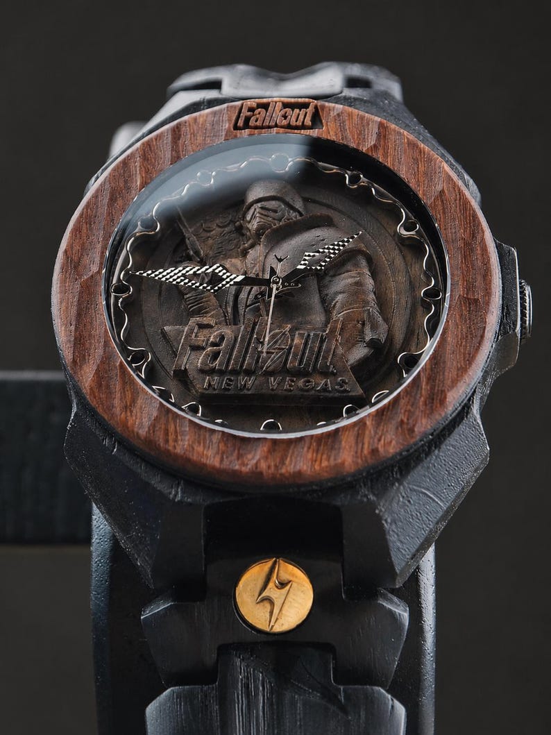 May include: A black wooden wristwatch featuring a dark wood bezel and a dial with a carved Fallout New Vegas character relief. The watch face displays the Fallout logo and text, with metallic hands and a small gold circular emblem on the strap.