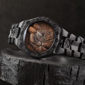 May include: A black wooden wristwatch featuring a dark, textured link band and a circular bezel with intricate Celtic knotwork engravings. The watch face displays a detailed brown wood grain illustration of two figures embracing, with silver hands.