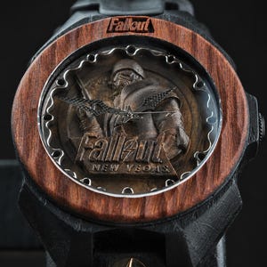 May include: A wristwatch featuring a dark wood bezel with the word Fallout engraved at the top. The watch face displays a relief of a character in armour with the text Fallout New Vegas. The watch has black hands and a black band with a gold bolt icon.