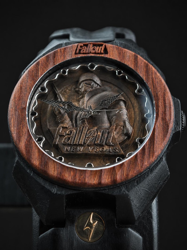 May include: A wristwatch featuring a dark wood bezel with the word Fallout engraved at the top. The watch face displays a relief of a character in armour with the text Fallout New Vegas. The watch has black hands and a black band with a gold bolt icon.