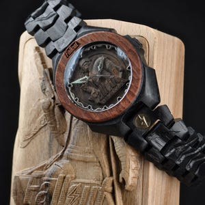 May include: A wooden wristwatch featuring a dark black link band and a reddish-brown circular bezel. The watch face displays the Fallout New Vegas logo and character imagery. It is presented on a carved wooden display stand with the Fallout New Vegas logo.