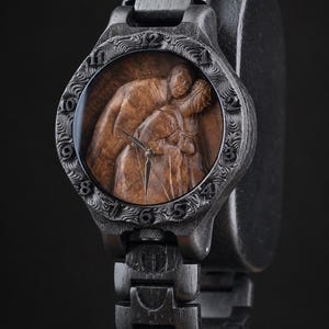 May include: A dark wood wristwatch featuring a carved, textured bezel with numerical hour markers. The watch face displays a detailed, three-dimensional brown wood carving of two figures embracing, with silver-toned hour and minute hands.