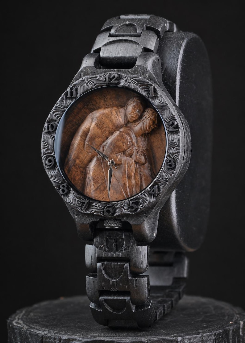 May include: A dark wood wristwatch featuring a carved, textured bezel with numerical hour markers. The watch face displays a detailed, three-dimensional brown wood carving of two figures embracing, with silver-toned hour and minute hands.