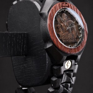 May include: A black wooden wristwatch featuring a reddish-brown wood bezel and a circular face with a Fallout New Vegas character engraving. The watch includes a black link band, a metallic lightning bolt emblem, and the Fallout logo on the dial.