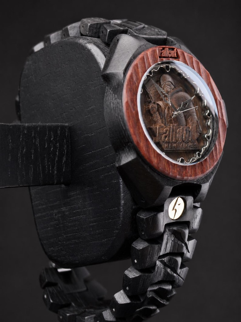 May include: A black wooden wristwatch featuring a reddish-brown wood bezel and a circular face with a Fallout New Vegas character engraving. The watch includes a black link band, a metallic lightning bolt emblem, and the Fallout logo on the dial.