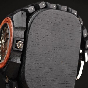 May include: A wristwatch featuring a dark black wooden band and case with a reddish-brown wooden bezel. The side of the case displays a small circular crown with a gold-coloured bull skull logo. The watch is mounted on a black wooden display block.