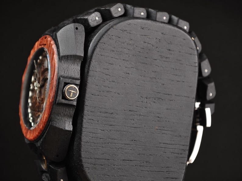 May include: A wristwatch featuring a dark black wooden band and case with a reddish-brown wooden bezel. The side of the case displays a small circular crown with a gold-coloured bull skull logo. The watch is mounted on a black wooden display block.