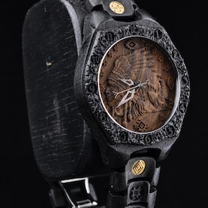Puede incluir: Un reloj de pulsera de madera negra con un bisel texturizado con números grabados y una esfera de madera marrón que muestra el perfil tallado de una persona con tocado. La correa incluye eslabones de madera negra con detalles metálicos dorados.