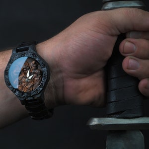 May include: A black wristwatch with a detailed, carved wooden face featuring a warrior figure and a silver emblem. The watch is worn on a wrist, and the image includes a sword.