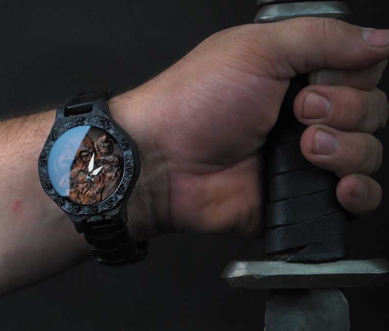 May include: A black wristwatch with a detailed, carved wooden face featuring a warrior figure and a silver emblem. The watch is worn on a wrist, and the image includes a sword.