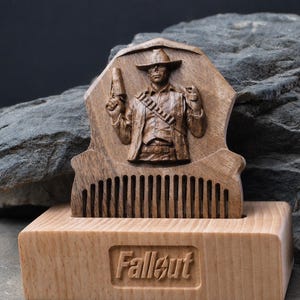 May include: A dark brown wooden comb featuring a carved relief of a cowboy figure holding a pistol, set into a light-coloured rectangular wooden base. The base has the word Fallout engraved in the centre of its front face in a recessed rectangular area.