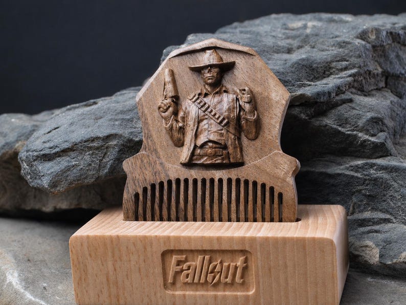 May include: A dark brown wooden comb featuring a carved relief of a cowboy figure holding a pistol, set into a light-coloured rectangular wooden base. The base has the word Fallout engraved in the centre of its front face in a recessed rectangular area.