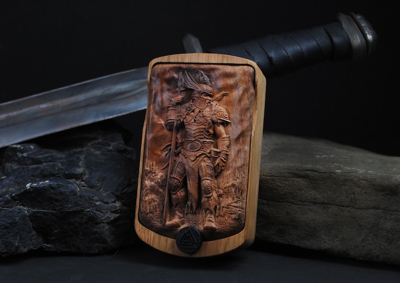 May include: A wooden carving of a warrior in full armor, holding a spear and a flag, set against a detailed landscape. The carving is mounted on a wooden frame. A sword with a leather-wrapped handle is in the background.