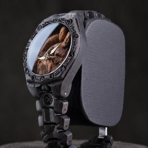 May include: A dark charcoal-colored wristwatch featuring a carved wooden face depicting two figures in an embrace. The watch has a textured, segmented band and a bezel engraved with numerical hour markers, complemented by thin, metallic hands.