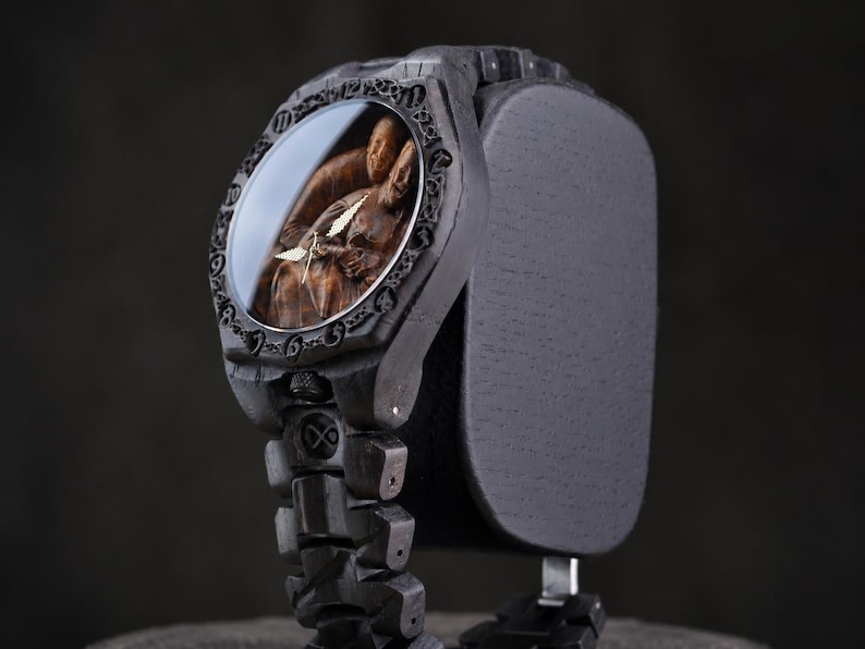May include: A dark charcoal-colored wristwatch featuring a carved wooden face depicting two figures in an embrace. The watch has a textured, segmented band and a bezel engraved with numerical hour markers, complemented by thin, metallic hands.