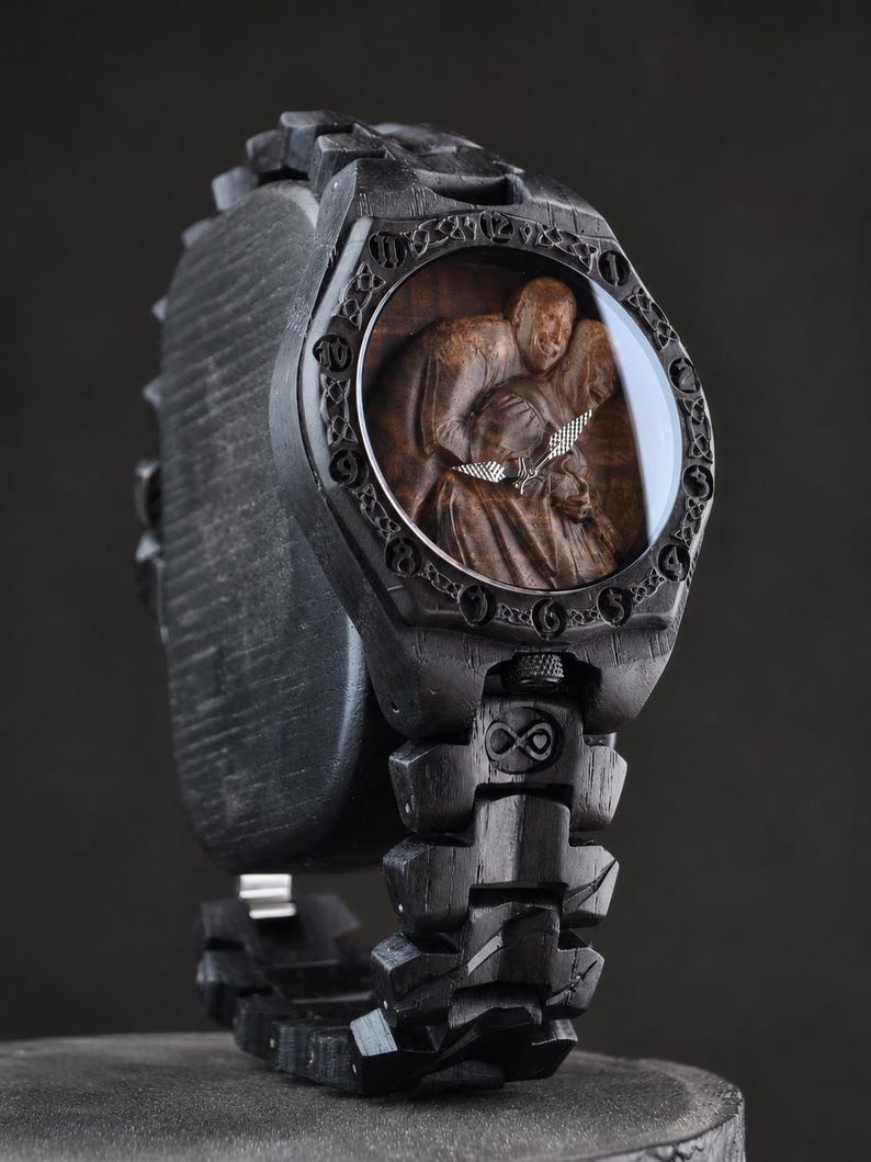 May include: A black wooden wristwatch featuring a carved relief of three figures on the dial. The bezel is engraved with circular symbols, and the link bracelet includes an infinity symbol. Silver-toned hands indicate the time on the dark wood face.