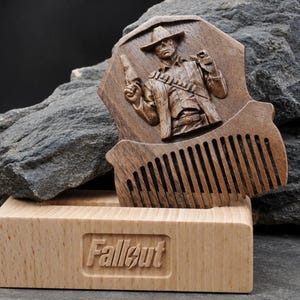 May include: A dark brown wooden comb featuring a carved relief of a cowboy figure holding a pistol, resting in a light-coloured wooden stand. The stand has the word Fallout engraved on the front in a rectangular recessed area with a lightning bolt.