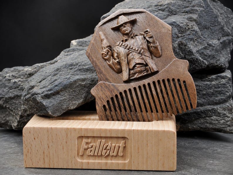 May include: A dark brown wooden comb featuring a carved relief of a cowboy figure holding a pistol, resting in a light-coloured wooden stand. The stand has the word Fallout engraved on the front in a rectangular recessed area with a lightning bolt.
