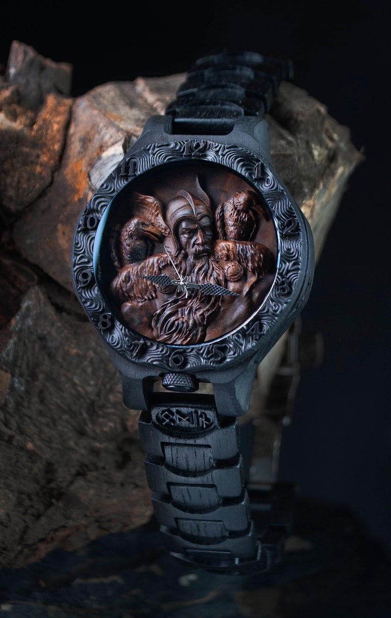 May include: A black wristwatch with a detailed, carved wooden face featuring a bearded figure and two birds. The watch has a black band and a decorative bezel. The watch face is brown and the hands are gold.