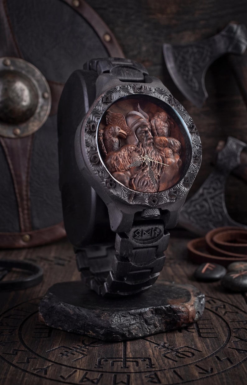 May include: A black wristwatch with a detailed, carved face depicting a Viking figure with birds. The watch has a dark, textured band and a decorative bezel. The watch is displayed on a black stand, with axes and shields in the background.