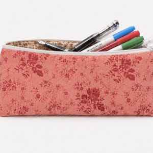 May include: A rectangular pencil case in a coral-pink fabric with a dark red floral pattern. The case features a white zipper closure, a gold-toned metal lobster clasp on the side, and a beige interior lining filled with several pens and markers.