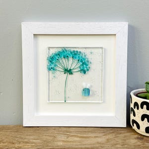 May include: A framed glass art piece featuring a teal flower with a green stem and a small bee. The artwork is set against a white background and framed in a white square frame. The piece is displayed on a wooden surface.