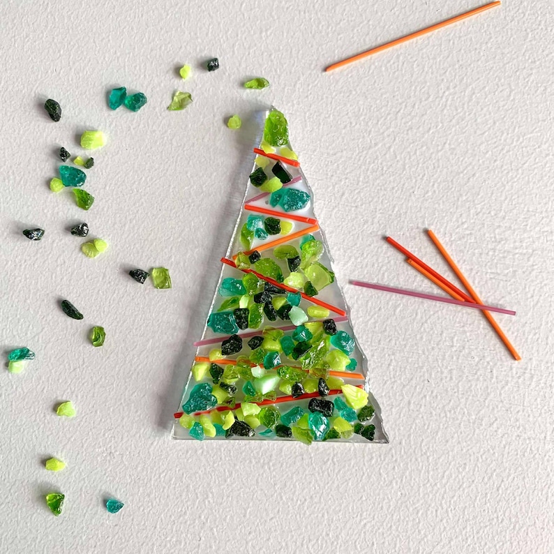 May include: A handcrafted Christmas tree made of clear glass, filled with green and black glass pieces. The tree is decorated with red and orange lines. Scattered around the tree are loose green glass pieces and orange sticks.