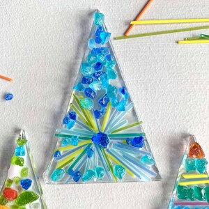 May include: Triangular glass art pieces with a Christmas tree design. The trees are decorated with blue and green glass pieces and lines. The background is white.