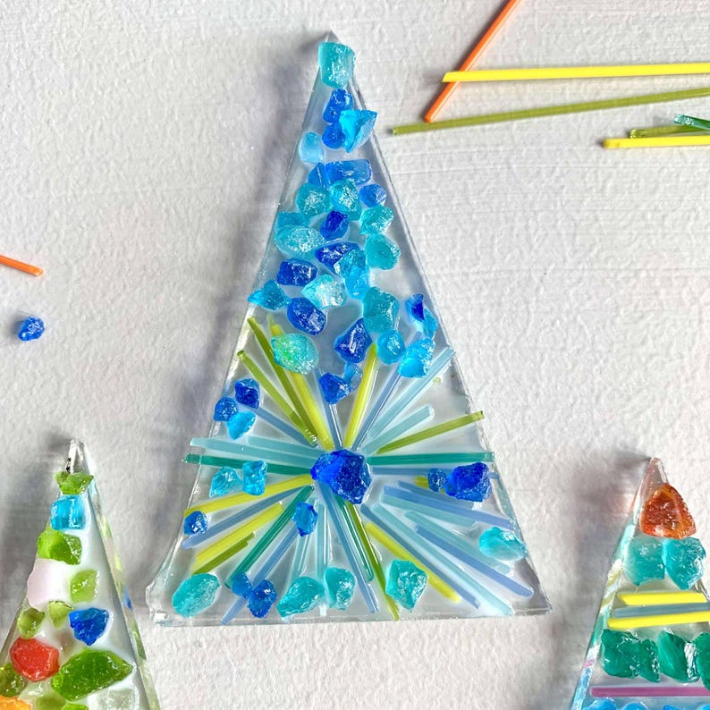 May include: Triangular glass art pieces with a Christmas tree design. The trees are decorated with blue and green glass pieces and lines. The background is white.