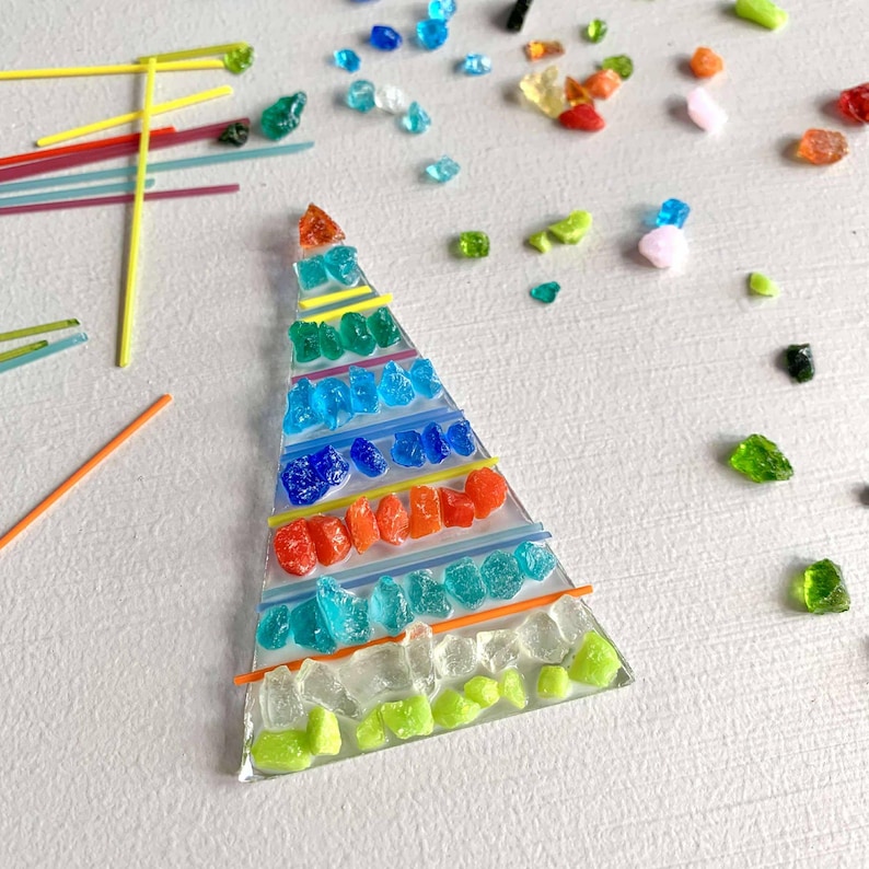 May include: A colorful, triangular glass art piece with layers of red, orange, blue, green, and clear glass pieces. Thin yellow and orange lines separate the layers. Scattered glass pieces in various colors surround the art piece.