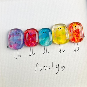 May include: A framed artwork featuring five colorful, abstract figures with drawn legs, arranged in a row. Each figure is a different color: purple, red, teal, yellow, and orange. The word "family" is handwritten below the figures.