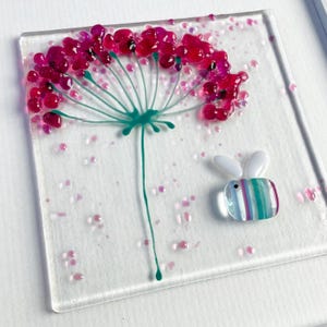 May include: A square glass art piece featuring a stylized flower with magenta petals and a teal stem. A small, colorful bee with white wings is next to the flower. Pink glass beads are scattered across the clear background.