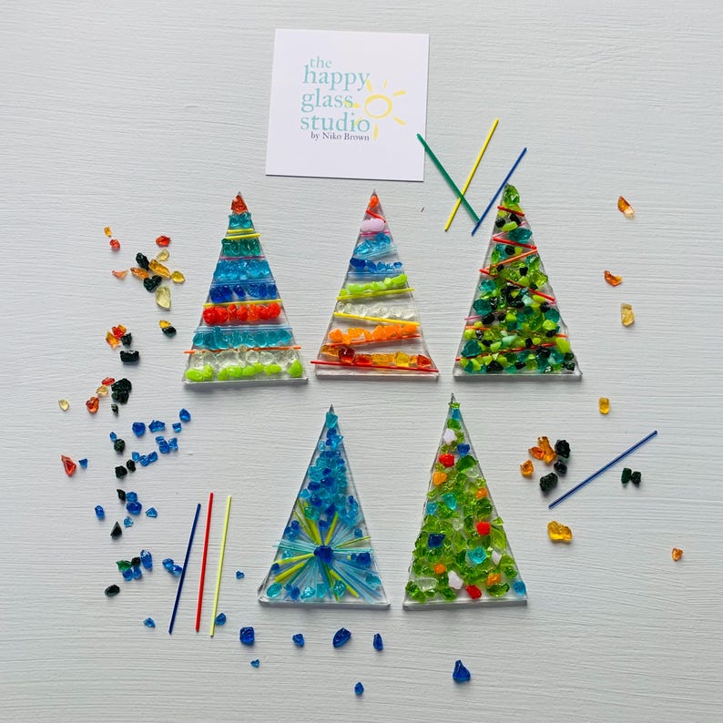 May include: Five colorful glass Christmas tree ornaments. Each tree is a different color and design, with various colored glass pieces and lines. The Happy Glass Studio logo is in the top left corner.