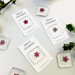 Könnte beinhalten: Weihnachtliche Glasornamente mit roten Stern- und Schneeflocken-Designs, präsentiert auf weißen Karten mit der Aufschrift "Merry Christmas". Die quadratischen Ornamente haben ein klares, strukturiertes Aussehen. Die Website-Adresse "www.nikobrown.co.uk" ist auf den Karten aufgedruckt.