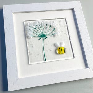 May include: A framed glass art piece featuring a dandelion with a green stem and white seed head, and a yellow and black bee. The artwork is set against a white background and framed in a white wooden frame.