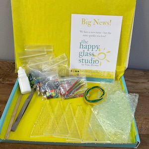 May include: Open box kit from The Happy Glass Studio, containing glass pieces, small bottles, tools, and colorful embellishments. The kit includes a card that reads "Big News!" and "We have a new name - but the same sparkle you love!"
