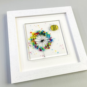 May include: A framed glass art piece featuring a colorful dandelion design with a black stem and a yellow and black bee. The dandelion is composed of colorful dots in shades of yellow, blue, red, and green. The frame is white.
