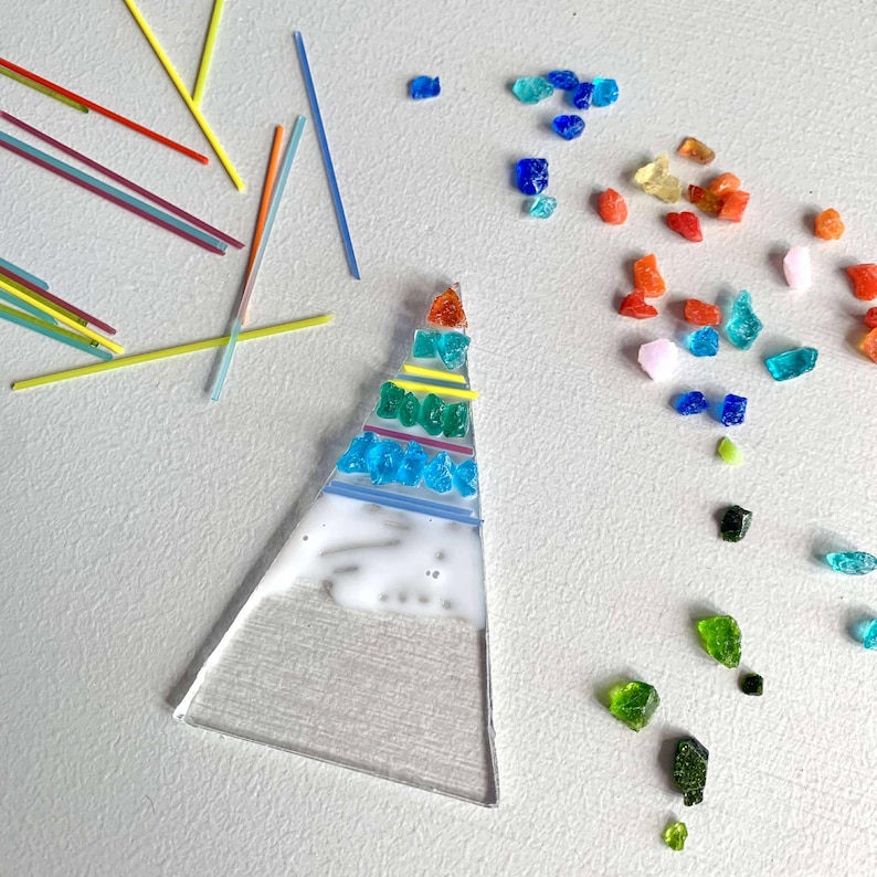 May include: A clear glass triangle with colorful glass pieces and lines arranged on its surface. The glass pieces are in shades of blue, green, orange, and red. Scattered around are small glass pieces and thin, colorful rods.