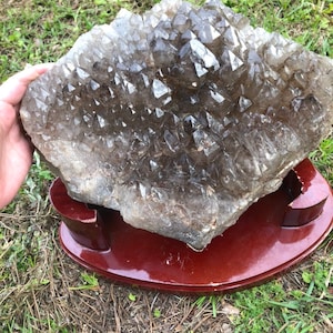 May include: A large, clear quartz crystal cluster with a smoky brown hue. The crystal is displayed on a dark brown wooden stand.