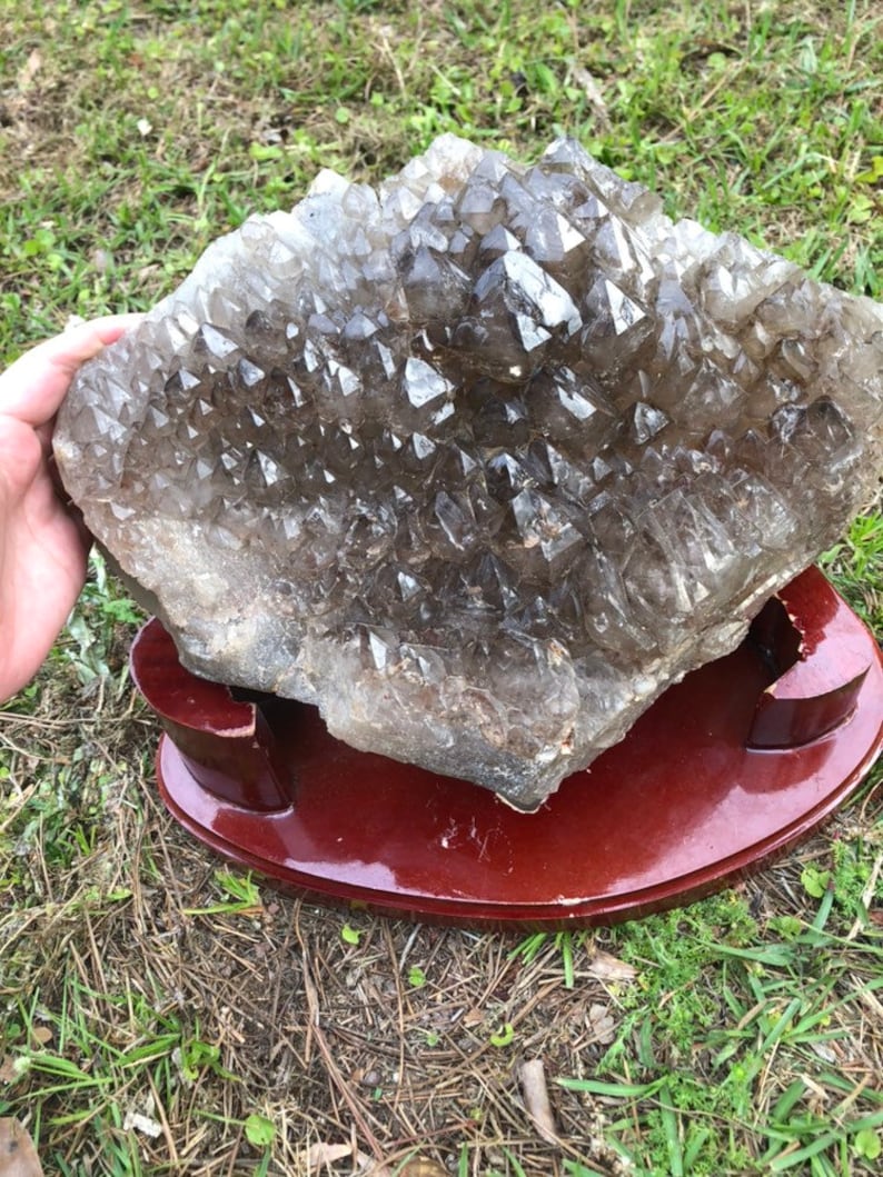 May include: A large, clear quartz crystal cluster with a smoky brown hue. The crystal is displayed on a dark brown wooden stand.